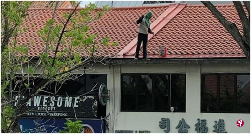 男子一手握刷子，一手提油漆桶，在两层楼店屋的屋顶上来回走动粉刷，对面顾客看得胆战心惊。（受访者提供）